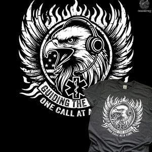 May include: Dark gray t-shirt featuring a distressed white graphic of an eagle wearing a headset, with flames and wings. The text reads "Guiding the Brave, One Call at a Time". The design includes an American flag and an emergency medical services symbol.