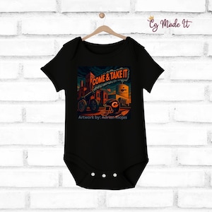 May include: Black baby bodysuit with a colorful graphic print. The design features a cannon, a football helmet, and the words "COME & TAKE IT." The artwork is signed by Adrian Riojas. The bodysuit is hanging on a wooden hanger against a white brick wall.