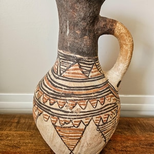 May include: A handmade, antique-style pitcher with a wide base and a handle. The pitcher features a dark brown neck and handle, with a cream-colored body adorned with geometric patterns in brown and black. The pitcher is likely made of clay.