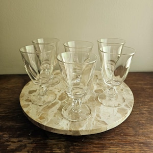 May include: Six clear, stemmed glasses with a textured design are arranged on a round, marble-patterned tray. The glasses have a classic, elegant shape, perfect for serving beverages. The tray sits on a dark wooden surface, creating a contrast with the light-colored tray and glasses.