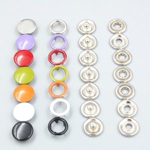 May include: Assortment of colorful and silver snap fasteners. The fasteners are arranged in rows, with the top row showing a silver, purple, red, green, orange, white, and black snap. The second row shows silver, purple, red, green, orange, white, and black snap rings. The third row shows silver snap studs.