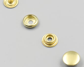 May include: Four gold-colored metal snap fasteners. The fasteners are in different stages of assembly. The first fastener is a cap with a small hole in the center. The second fastener is a base with a small hole in the center. The third fastener is a base with a small hole in the center and a decorative ring around the edge. The fourth fastener is a dome-shaped cap.