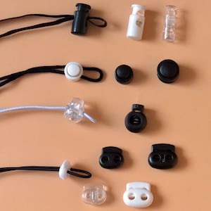 Può includere: Assortimento di blocca-corda e toggle in plastica nera e bianca. I blocca-corda sono rotondi e hanno un foro al centro. I toggle sono rettangolari e hanno due fori.