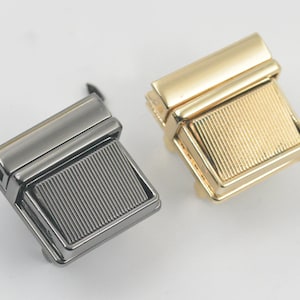 May include: Two metal clasps, one gold and one silver, with a ridged design. The clasps are square shaped and have a hinged design.