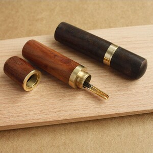 May include: Two wooden needle cases with brass accents. One case is open and reveals a set of needles. The cases are made of dark wood and light wood.
