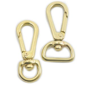Puede incluir: Dos cierres giratorios de metal dorado con un anillo en D en un lado. Los cierres se utilizan comúnmente para sujetar correas o correas a bolsos, carteras u otros artículos.