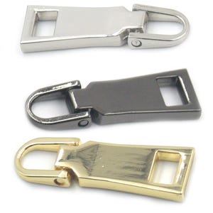 Op de afbeelding: Drie metalen ritstrekkers in zilver, gunmetal en goud. Elke trekker heeft een rechthoekig lichaam met een vierkante opening en een gebogen, U-vormige lus voor bevestiging. De trekkers zijn ontworpen voor gebruik op ritsen.