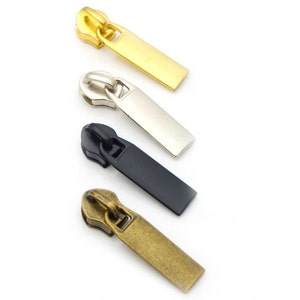 Op de afbeelding: Vier metalen ritstrekkers in goud, zilver, zwart en antiek messing. Elke trekker heeft een rechthoekig lipje voor gemakkelijk vastpakken. De ritstrekkers zijn diagonaal geplaatst tegen een witte achtergrond, waarbij hun verschillende afwerkingen worden getoond.