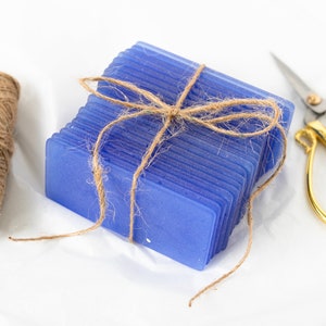 May include: A stack of blue rectangular tiles tied together with twine. The tiles are arranged in a neat stack and are tied with a piece of twine. There is a spool of twine and a pair of gold scissors on the white surface.