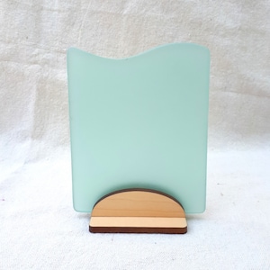 May include: A frosted glass sign with a wavy top edge, standing on a wooden base. The sign is light green and the base is a light brown colour.