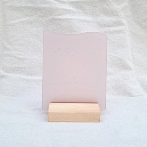 Peut inclure: Un rectangle de verre rose clair avec un bord supérieur ondulé, monté sur un petit socle en bois naturel. Le verre a un aspect légèrement givré et est présenté sur un fond de tissu blanc.