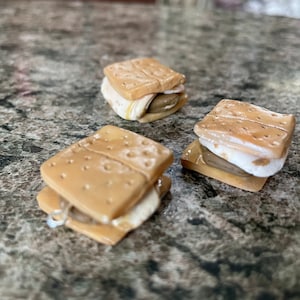 以下が含まれることがあります： 茶色のグラハムクラッカー、白いマシュマロ、茶色のチョコレートでできたミニスモアが3つ。スモアは灰色の斑点のある表面に並べてあります。