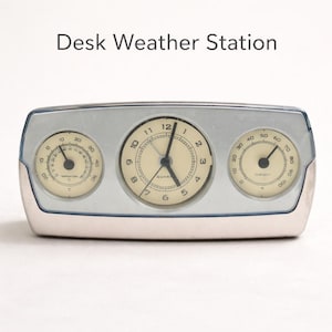 Puede incluir: Estación meteorológica de escritorio plateada con tres esferas circulares. La esfera central es un reloj, flanqueado por un termómetro a la izquierda y un higrómetro a la derecha. El texto "Desk Weather Station" está arriba.