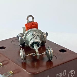 以下が含まれることがあります： 金属コネクタ付きの茶色の長方形ベース、銀色のダイオード、オレンジ色のコンポーネントなど、電子部品のクローズアップ。ベースには「K401T 0160 UTK4」の文字が書かれています。