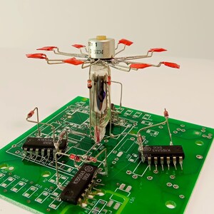 Puede incluir: Una placa de circuito verde con componentes electrónicos, incluidos circuitos integrados y un tubo de vacío central con detalles en rojo. Los componentes están dispuestos en la placa, creando un complejo ensamblaje electrónico.