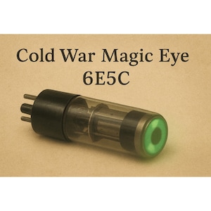 以下が含まれることがあります： 「Cold War Magic Eye 6E5C」の文字が入ったヴィンテージ真空管。 黒いベースと透明なガラスボディで、端には緑色の光る目があります。 背景は薄茶色です。