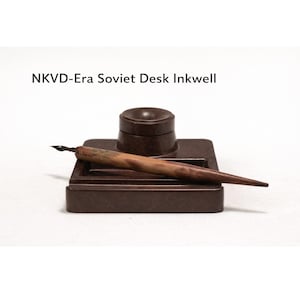 May include: A brown NKVD-Era Soviet desk inkwell with a matching pen. The inkwell has a square base with a circular ink pot and a pen rest. The pen is made of wood and has a metal nib. The text "NKVD-Era Soviet Desk Inkwell" is above the inkwell.