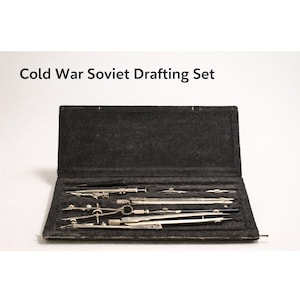 Pode incluir: Um conjunto de desenho soviético da Guerra Fria em uma caixa preta aberta. O conjunto inclui várias ferramentas de desenho prateadas, como compassos e divisores. O texto "Cold War Soviet Drafting Set" é exibido acima da caixa.