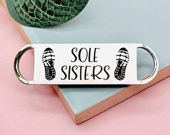 May include: A silver metal keychain with a black inscription that reads "SOLE SISTERS" and two black shoe prints.