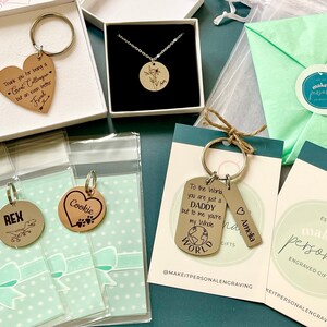 May include: Assortment of personalized engraved gifts, including keychains, necklaces, and a small bag. The keychains feature various designs, including a heart with the word "Cookie", a dog paw print, a dinosaur, and a world map. The necklace features a round pendant with a floral design. The bag is green and has a white circle with the text "make it personal" on it.