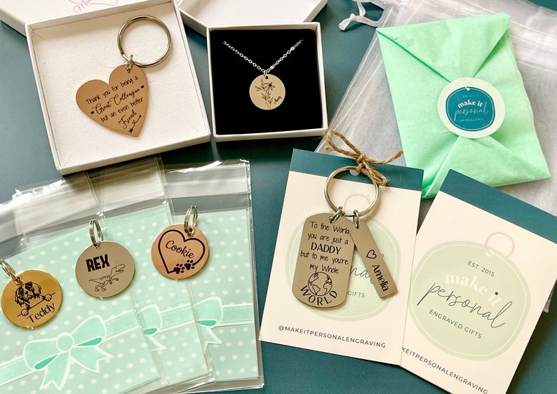 May include: Assortment of personalized engraved gifts, including keychains, necklaces, and a small bag. The keychains feature various designs, including a heart with the word "Cookie", a dog paw print, a dinosaur, and a world map. The necklace features a round pendant with a floral design. The bag is green and has a white circle with the text "make it personal" on it.