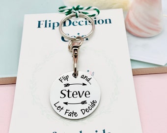 以下が含まれることがあります： 矢印が反対方向を向いている「Flip and... Steve Let Fate Decide」と書かれた丸いチャームが付いたシルバーのキーホルダー。キーホルダーは、「Let fate decide... @makeitpersonalengraving」というテキストが書かれた白いカードの上に置かれています。