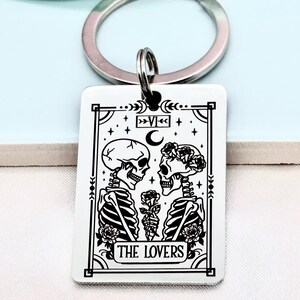May include: A rectangular keychain with a silver key ring. The white pendant features a black and white illustration of two skeletons holding a rose, with the text "THE LOVERS" at the bottom. The design is framed with decorative elements.