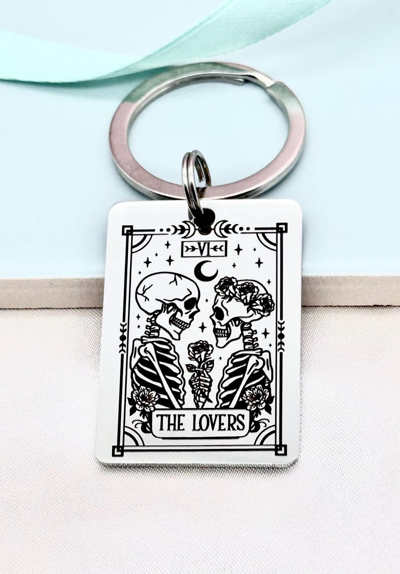 May include: A rectangular keychain with a silver key ring. The white pendant features a black and white illustration of two skeletons holding a rose, with the text "THE LOVERS" at the bottom. The design is framed with decorative elements.