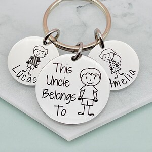 May include: Silver keychain with three round tags. The center tag has the text "This Uncle Belongs To" and a stick figure drawing of a boy. The other two tags have stick figure drawings of a boy and a girl and the names "Lucas" and "Amelia".