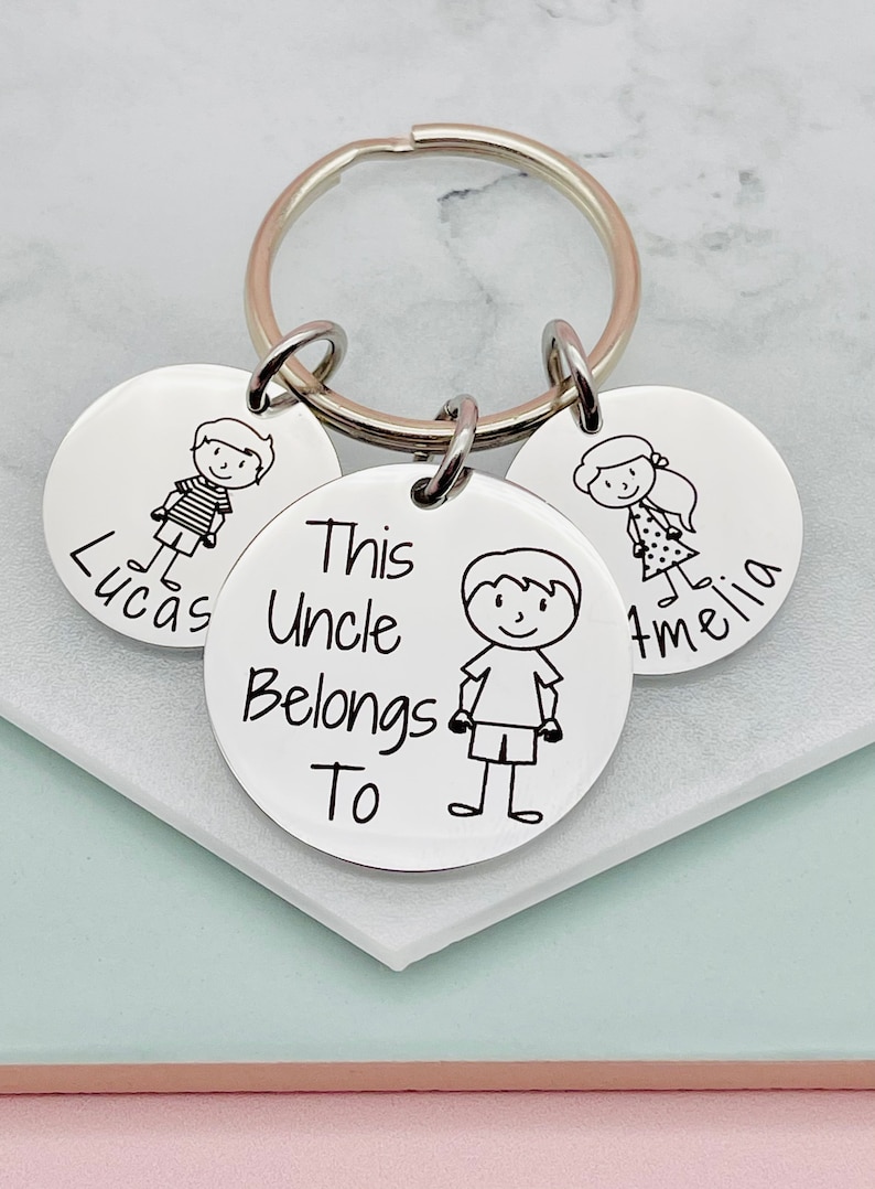 May include: Silver keychain with three round tags. The center tag has the text "This Uncle Belongs To" and a stick figure drawing of a boy. The other two tags have stick figure drawings of a boy and a girl and the names "Lucas" and "Amelia".