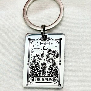 May include: Silver-colored keychain with a rectangular pendant featuring a black and white illustration of two skeletons facing each other, holding a rose. The text "THE LOVERS" is at the bottom. A silver ring is attached to the top.