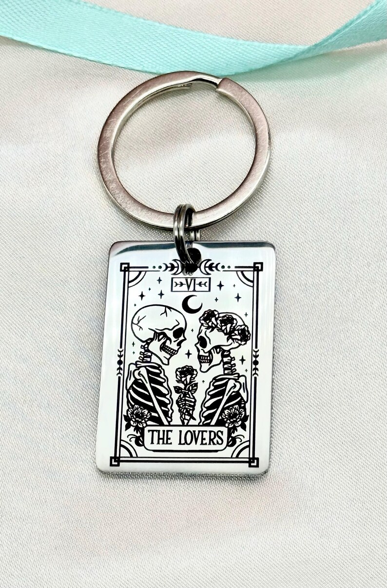 May include: Silver-colored keychain with a rectangular pendant featuring a black and white illustration of two skeletons facing each other, holding a rose. The text "THE LOVERS" is at the bottom. A silver ring is attached to the top.