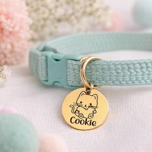 May include: A light blue cat collar with a gold-colored tag. The tag features a cartoon cat design and the name "Cookie" in a script font. The collar has a plastic buckle and a gold-colored metal ring.