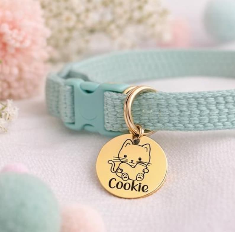 May include: A light blue cat collar with a gold-colored tag. The tag features a cartoon cat design and the name "Cookie" in a script font. The collar has a plastic buckle and a gold-colored metal ring.