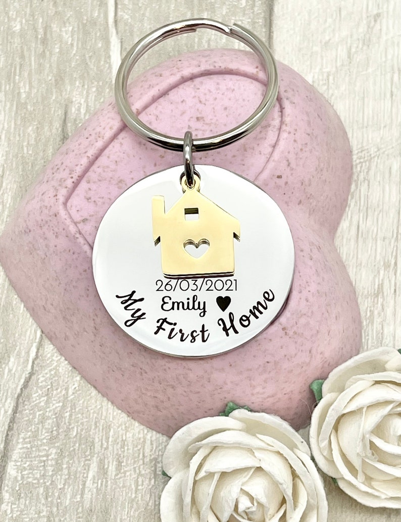 May include: A silver-colored keychain with a round pendant featuring a gold house silhouette with a heart cutout. The text "My First Home" and the date "26/03/2021" are printed on the pendant. The keychain is displayed with white roses.