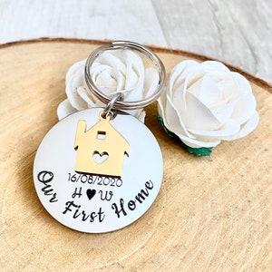 May include: A round, silver keychain with the words "Our First Home" in black script. A gold-colored house charm with a heart cutout is attached. The date "16/08/2020" and initials "H &hearts; W" are also present. White flowers are in the background.