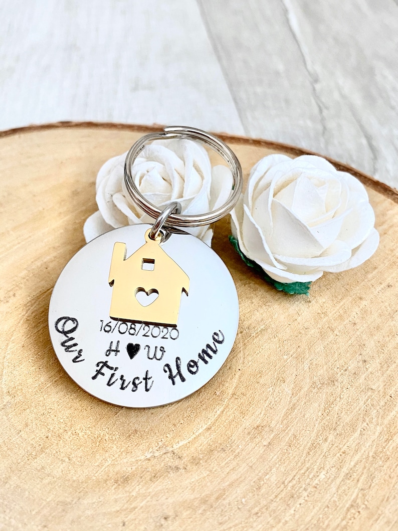 May include: A round, silver keychain with the words "Our First Home" in black script. A gold-colored house charm with a heart cutout is attached. The date "16/08/2020" and initials "H &hearts; W" are also present. White flowers are in the background.