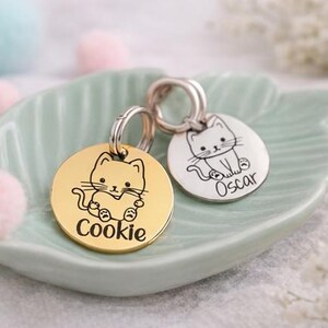 May include: Two round pet tags with cat illustrations. One is gold-colored with the name "Cookie", and the other is silver-colored with the name "Oscar". Both tags have a silver ring for attachment. The tags are on a light green, leaf-shaped dish.