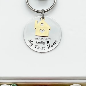 May include: A silver keychain with a gold house charm and a round tag. The tag is engraved with the words "My First Home", a date, and a name. The keychain is attached to a white background.