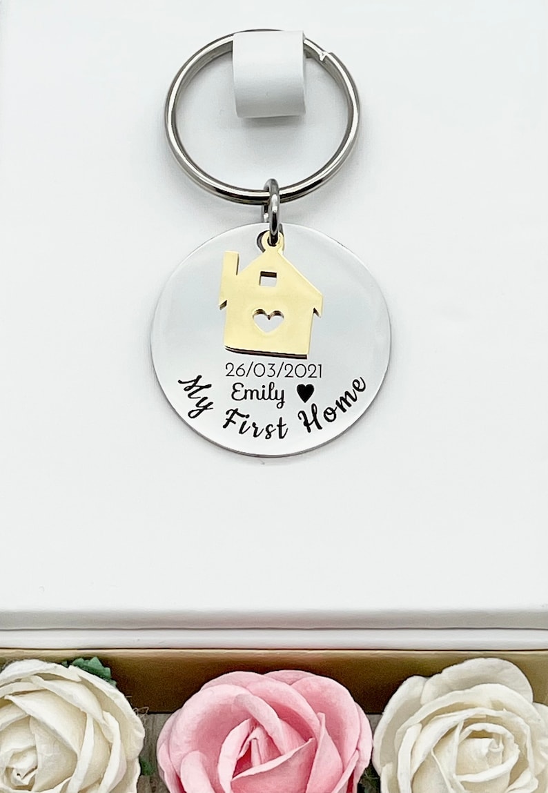 May include: A silver keychain with a gold house charm and a round tag. The tag is engraved with the words "My First Home", a date, and a name. The keychain is attached to a white background.