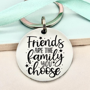 May include: A silver-colored round keychain with the text "Friends are the family you choose" in black script. The keychain has a silver ring and a light green ribbon.