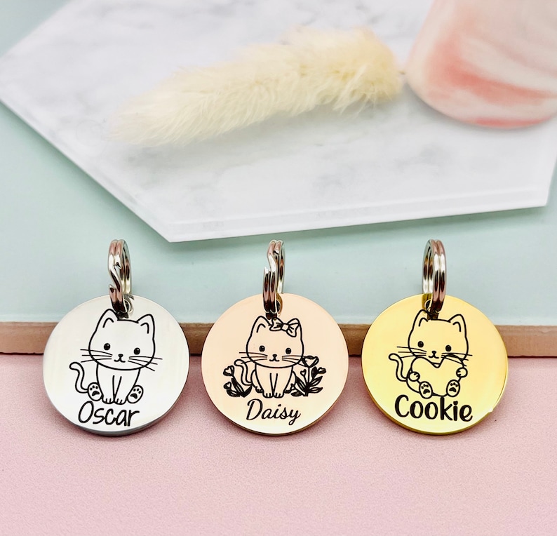 May include: Three round pet tags with cat illustrations and names. The tags are silver, gold, and rose gold. Each tag has a different cat design and name: Oscar, Daisy, and Cookie. The tags have a silver ring for attachment.