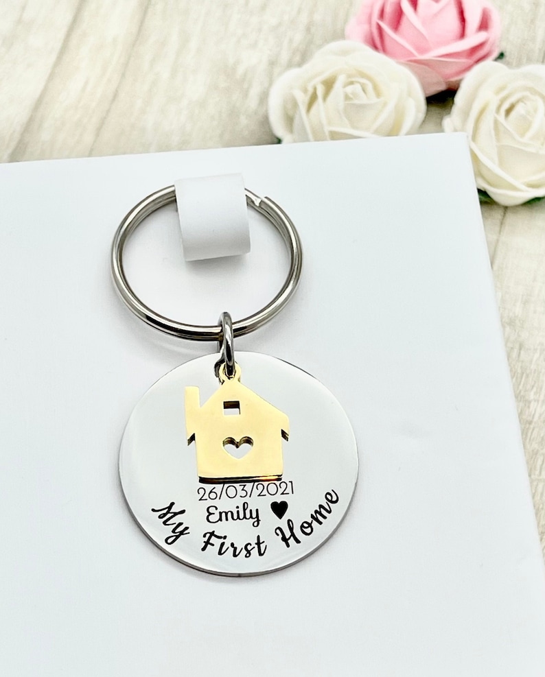 May include: A silver-colored round keychain with a gold-colored house charm. The keychain has the text "My First Home" and the name "Emily" with a date. The keychain is attached to a silver key ring.