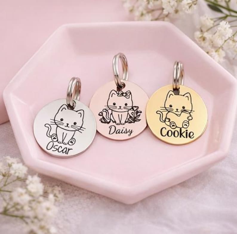 May include: Three circular metal pet ID tags with silver, rose gold, and gold finishes. Each tag features an engraved cat illustration and a name: Oscar, Daisy, or Cookie. Each tag includes a metal split ring for attachment to a collar.