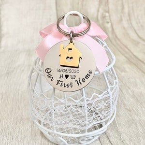 May include: A silver-colored keychain with a gold house charm and the words "Our First Home" engraved on it. The date "16/08/2020" is also included. A pink ribbon and a white decorative cage are in the background.