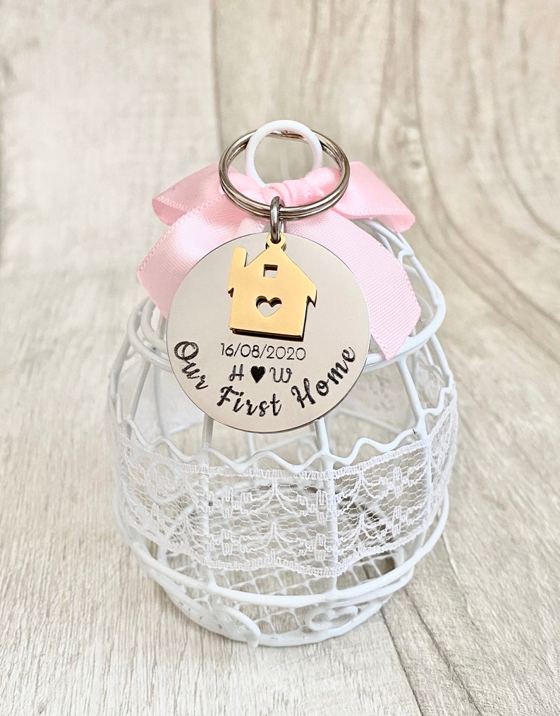 May include: A silver-colored keychain with a gold house charm and the words "Our First Home" engraved on it. The date "16/08/2020" is also included. A pink ribbon and a white decorative cage are in the background.
