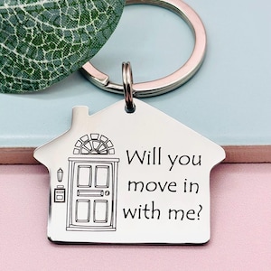 May include: A silver house-shaped keychain with a black outline of a door and the text "Will you move in with me?"