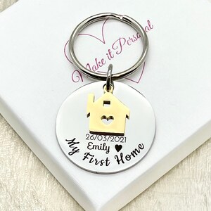 May include: A silver-colored round keychain with a gold-colored house charm. The keychain has the text "My First Home", the date "26/03/2021", and the name "Emily". The keychain is attached to a silver ring.