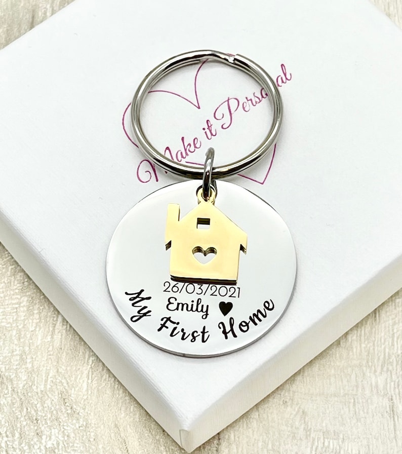 May include: A silver-colored round keychain with a gold-colored house charm. The keychain has the text "My First Home", the date "26/03/2021", and the name "Emily". The keychain is attached to a silver ring.