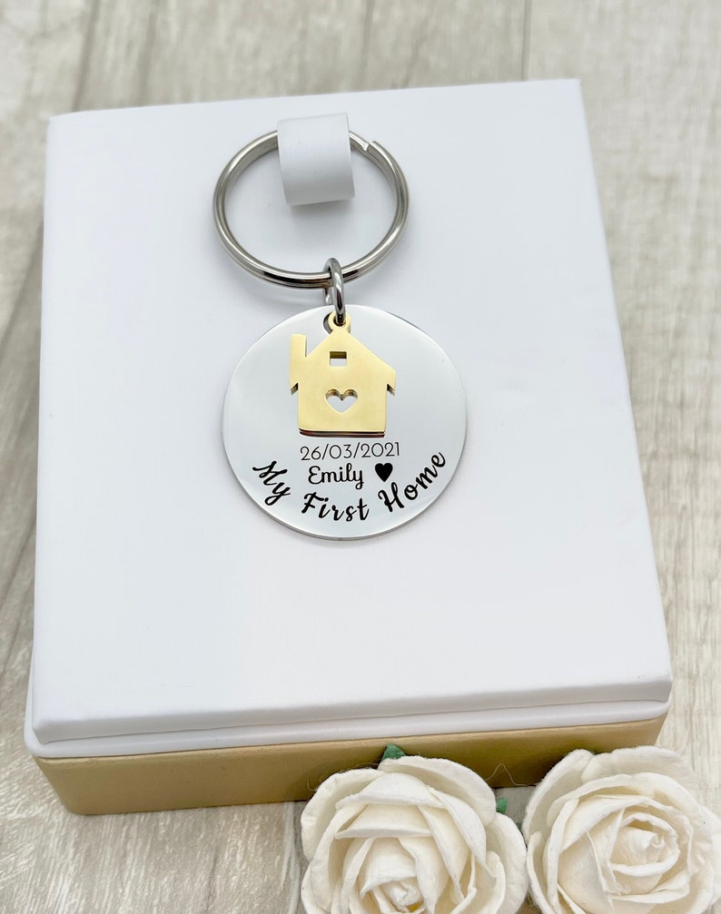 May include: A silver-colored keychain with a gold house charm and a key ring. The charm has a heart-shaped cutout and the text "My First Home" and the date "26/03/2021". The keychain is presented on a white box with a gold base.